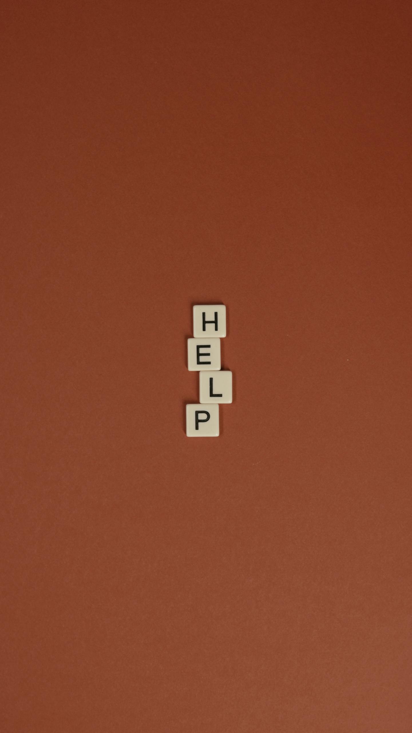 Vertical arrangement of Scrabble tiles spelling 'Help' on a brown background, minimalist composition.